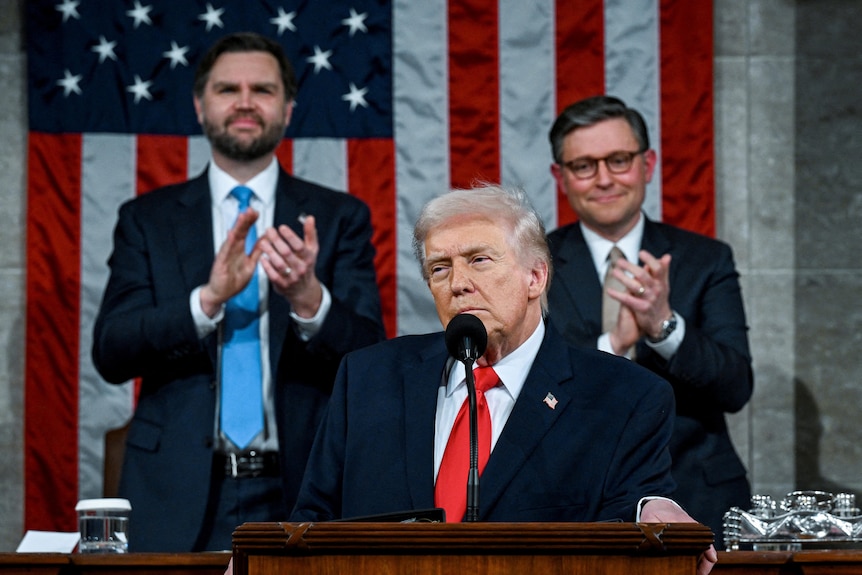 Three men in dark suits — Donald Trump with JD Vance and Mike Johnson — on a stage in front of an American flag.