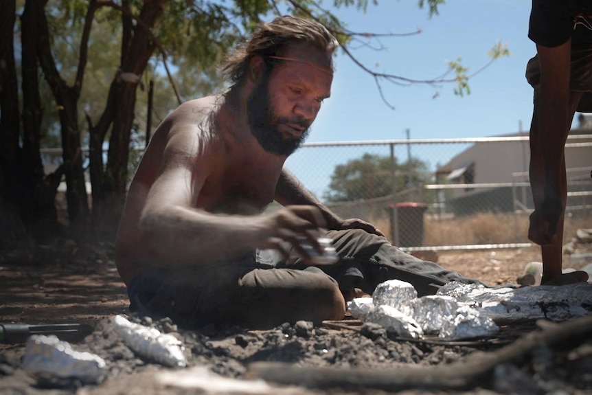 A man sits next to a campfire making a meal.