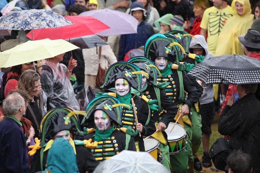 A troupe of drummers walk through a crowd of people with umbrellas