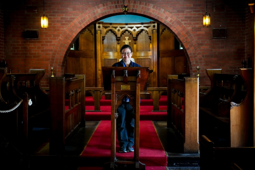 A dark haired woman with glasses stands behind a wooden pulpit on red carpet in a brick and wooden room with pews.