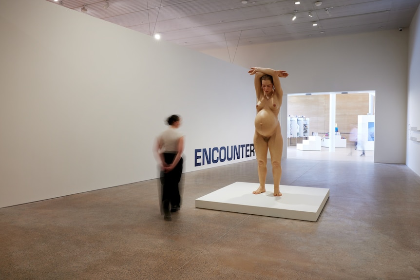 In a gallery space, a woman approaches a large hyperreal sculpture of a naked pregnant woman, arms stretched over her head.