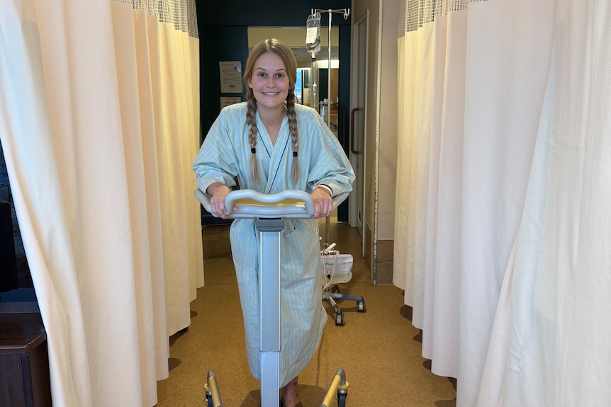 A woman in hospital robes walks with an assisted walking machine down a hospital hall with white curtains on either side.
