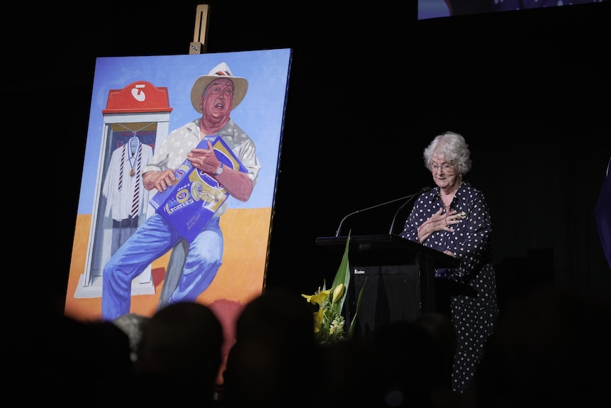 A woman speaking at a lectern on a stage, one hand on her chest, next to a large painting of a man playing a beer carton.