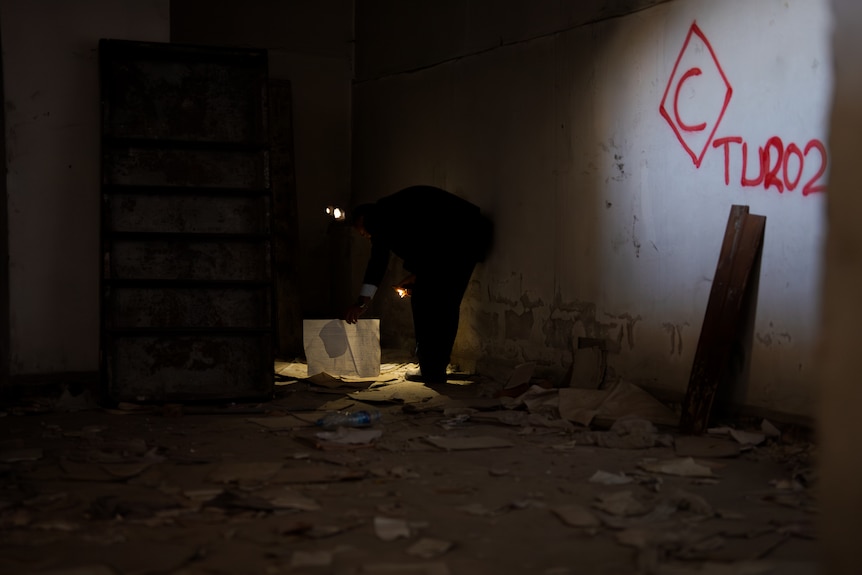 A man crouching in a dark room with a headlight searching for documents.