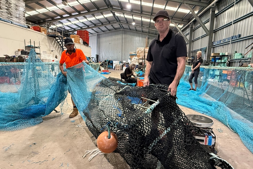 Two men hold up fishing nets as a woman stands behind them in a shed.