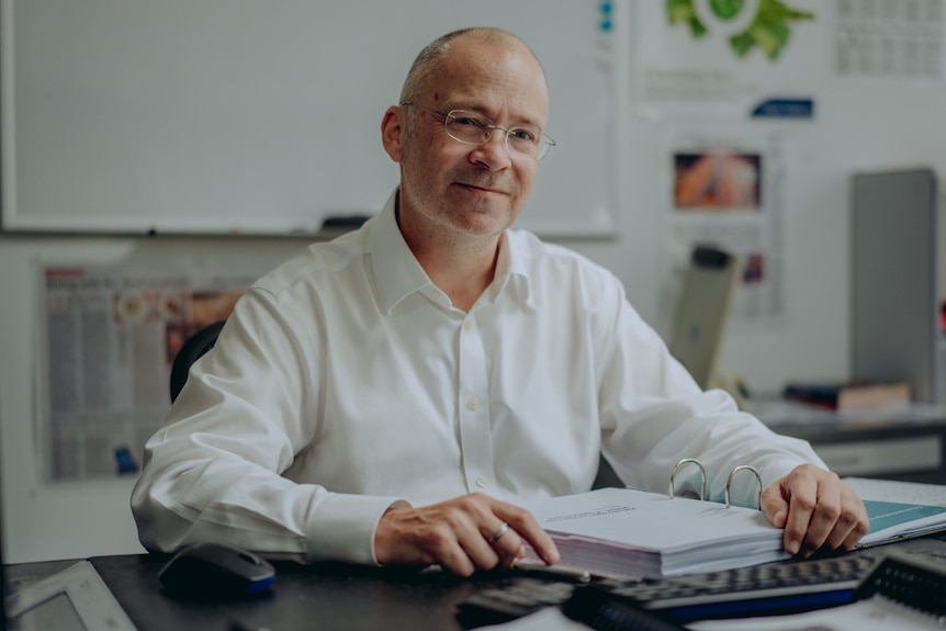 A man with no hair and wearing a white shirt sits at an office desk in front of a binder
