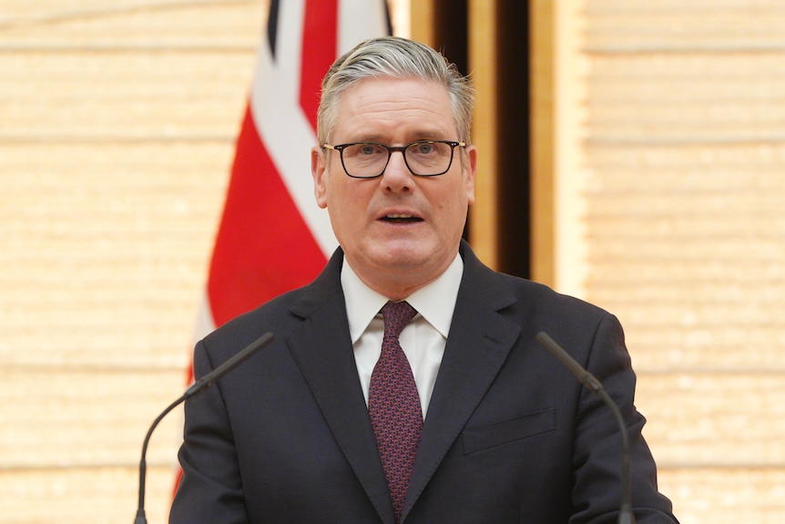 A middle aged caucasian man wearing a suit and glasses speaks into a mic
