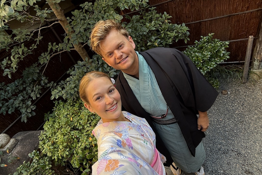 A man and woman in kimonos take a selfie in front of a bonsai.
