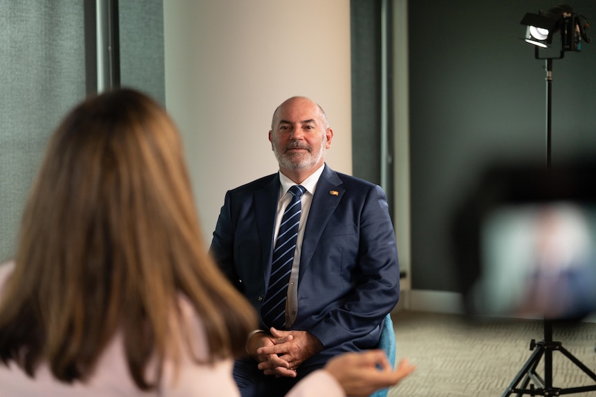 White man, navy blue suit, gray hair, sitting on chair in conversation with a woman, pink suit, brown hair in foreground.