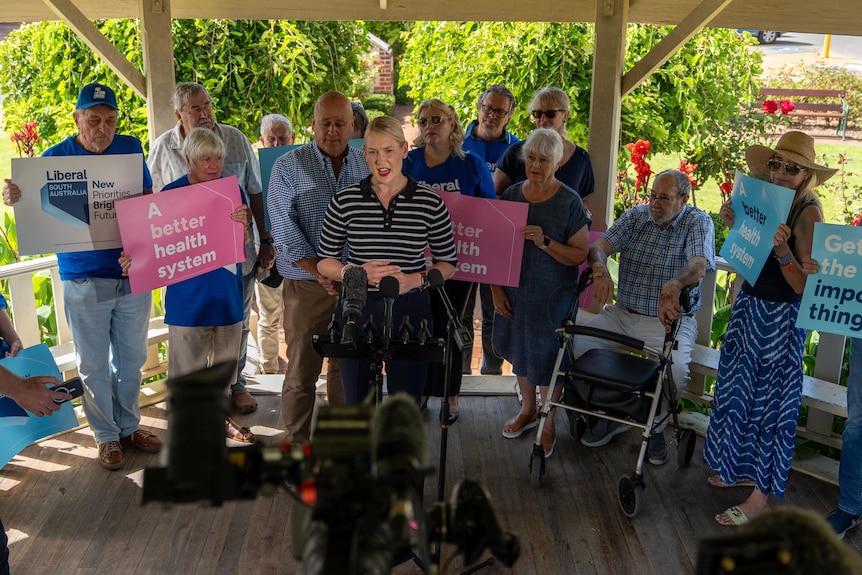 A woman speaks into multiple microphones while surrounded by a group of people.