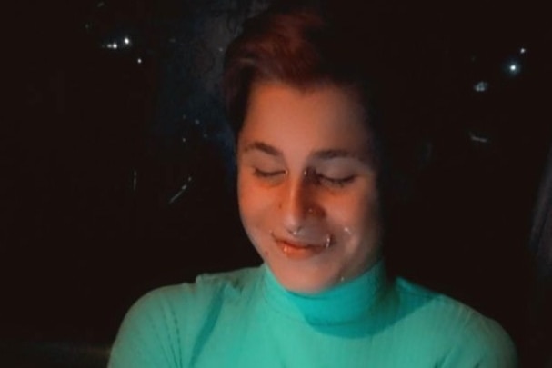 Image of Alexx who has short hair and facial piercings around their mouth and nose. They smile at their phone screen.