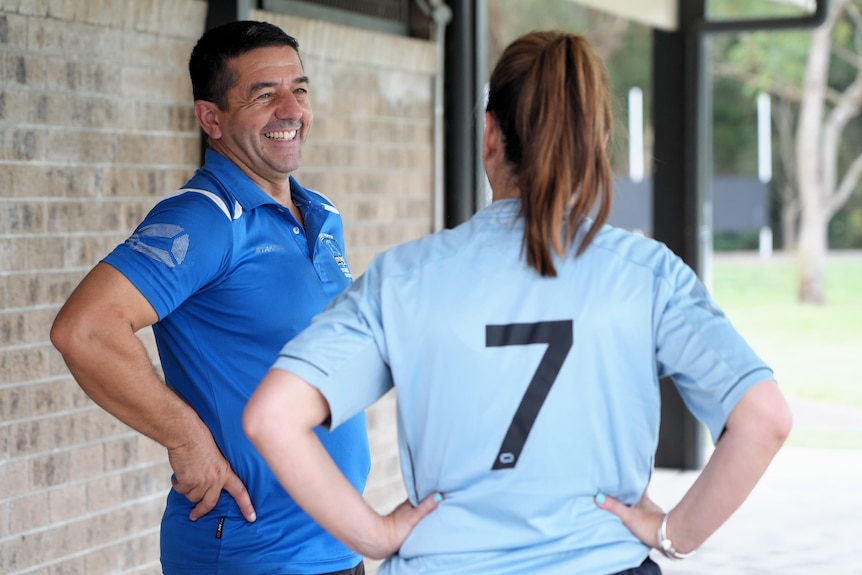 A man wearing a blue polo shirt has hands on hips and smiles as he speaks to a female footballer