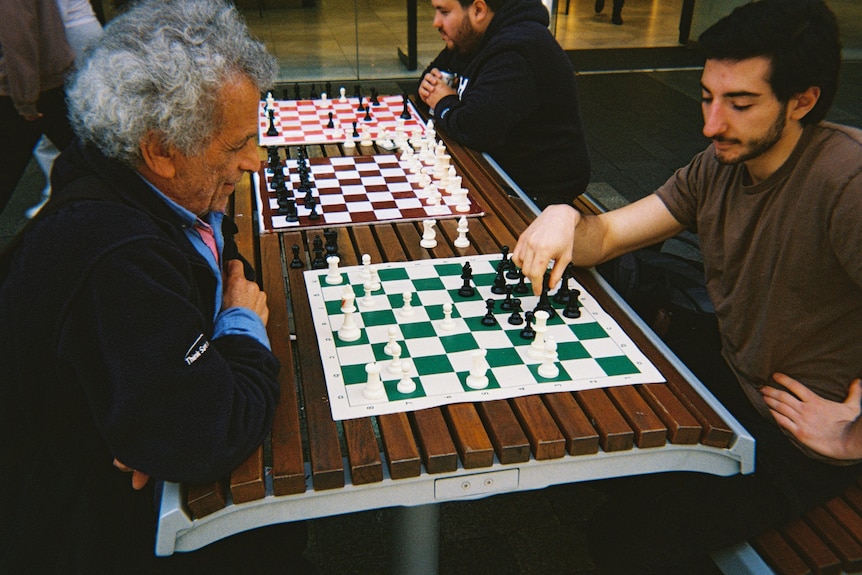 Two men play chess on an outdoor table, where others are playing in the background