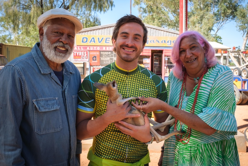 Three actors smiling at the camera, one is holding a baby kangaroo.