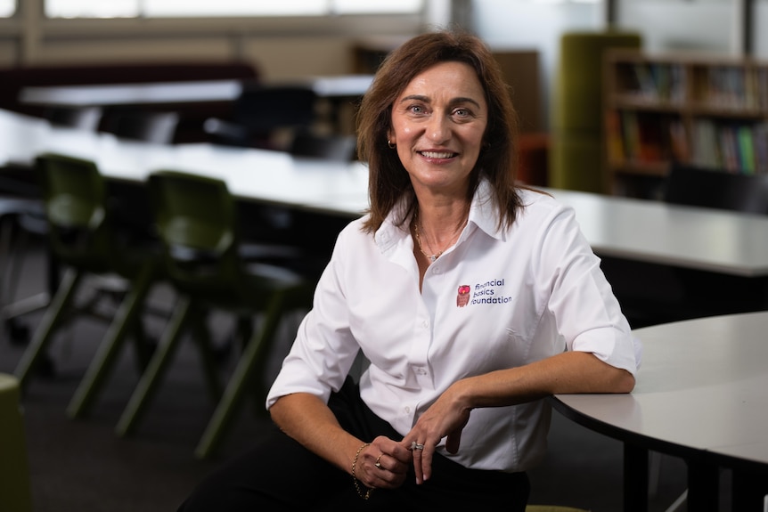A woman with straight brown hair, wearing a white mid-length-sleeved shirt, smiles, and rests her elbow on a desk.
