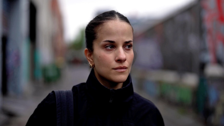 A close portrait of a woman with a neutral expression looking ahead. Out of focus behind her is a laneway with graffiti.