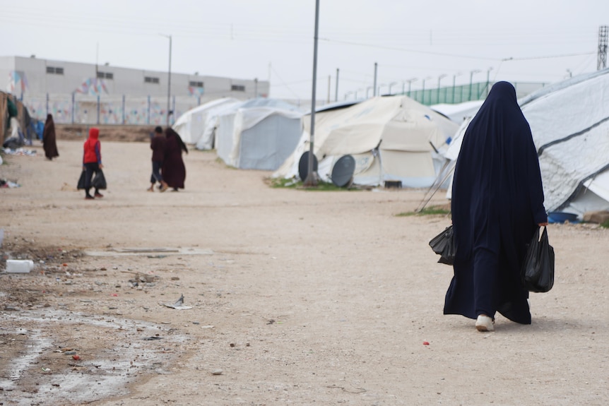 A woman in a black niqab walks along a row of tents, with children seen in the distance.