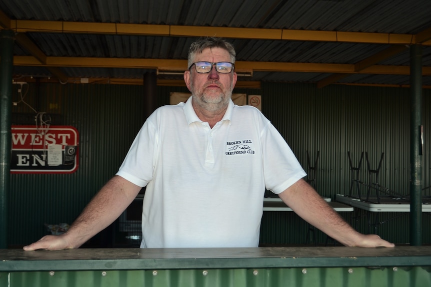 Regan wears a white shirt and glasses, leaning on a counter with his arms wide, staring out.