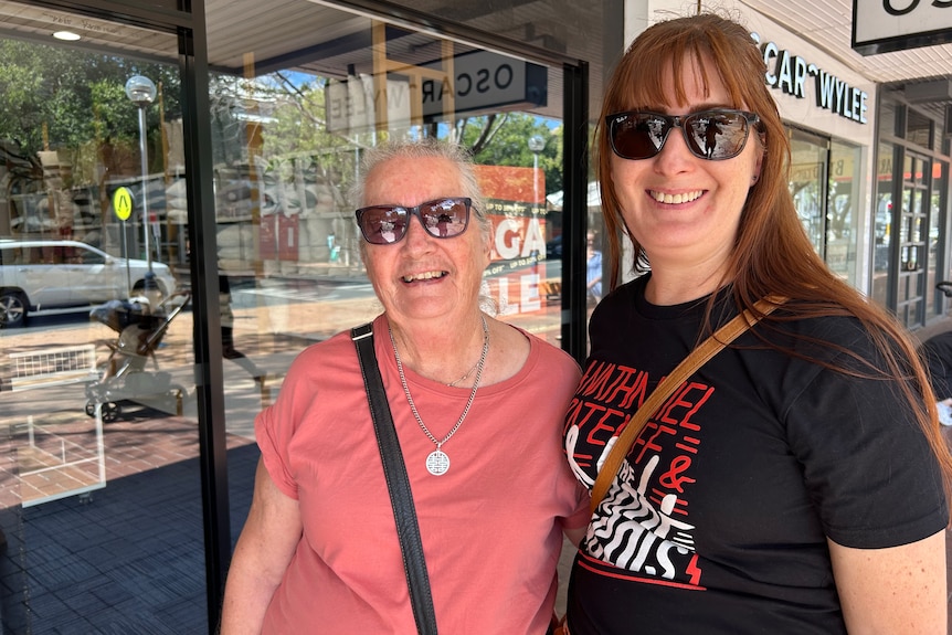 A young woman with brown hair and a black t shirt stands next to her mother in a pink shirt