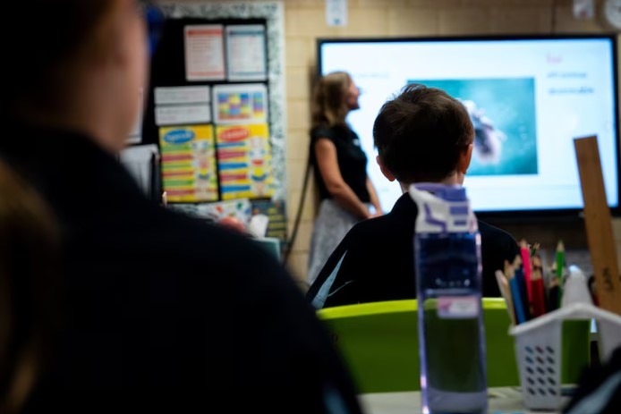 Unidentifiable students sitting at desks listen to a teacher in a primary school classroom.