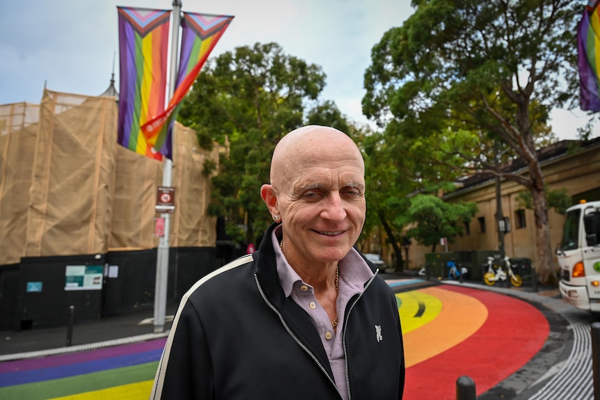 A smiling, older man in a dark tracksuit top on a city street with a rainbow marking on the road.