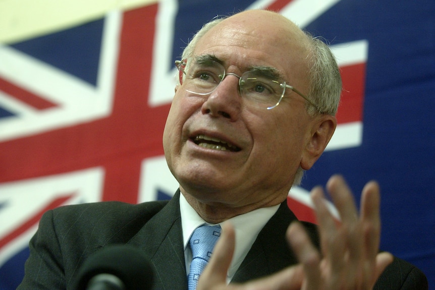 John Howard head and shoulders shot speaking while standing in front of an Australian flag. He wears a suit and tie
