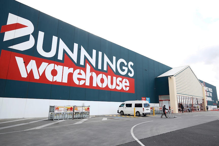 The exterior of a large Bunnings Warehouse store. A couple of cars are parked outside and a person is pushing a trolley.
