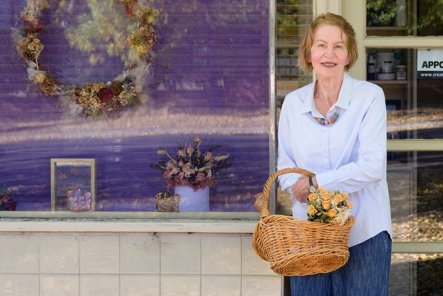 Woman stands outside shop holding basket with dried flowers in it.