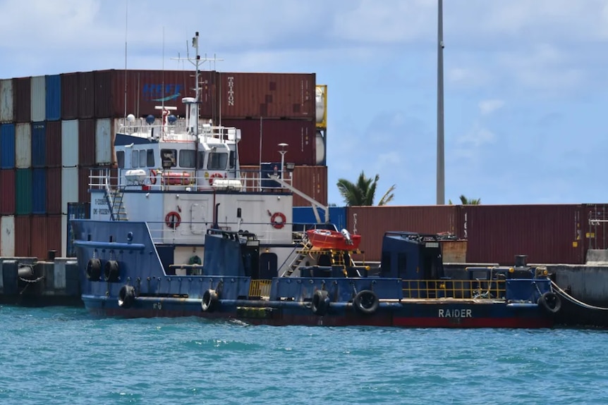 A trawler docked at a wharf stacked with shipping containers.