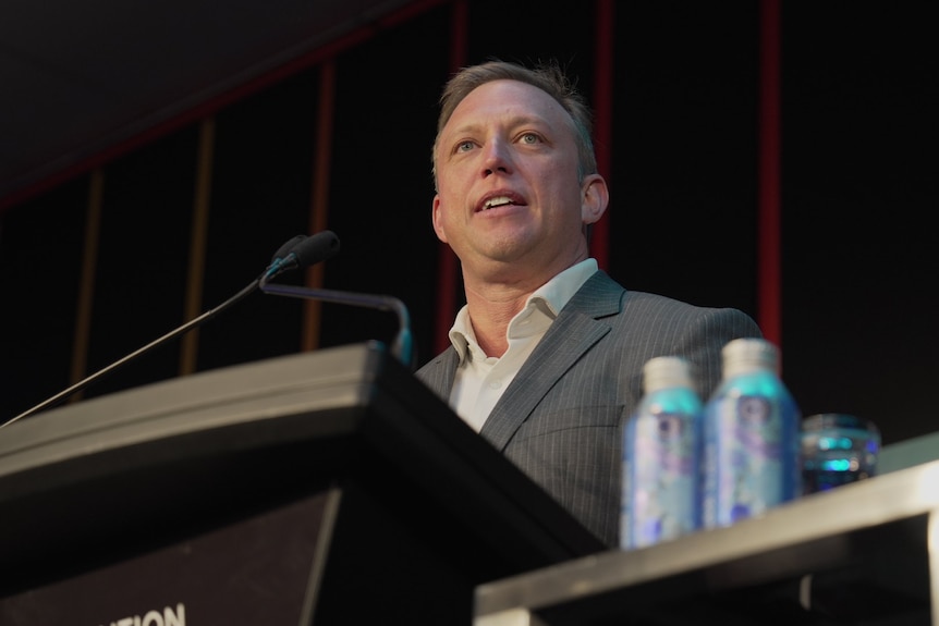 A man in aan open neck shirt and suit jacket speaking at a podium. 