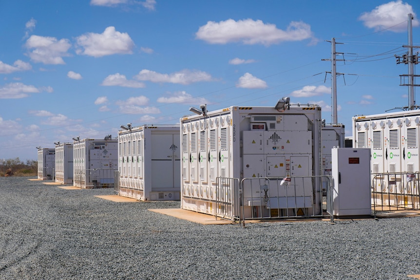 Rows of cubic machinery sits on a gravel industrial park underneath transmission line and a blue sky dotted with small clouds.