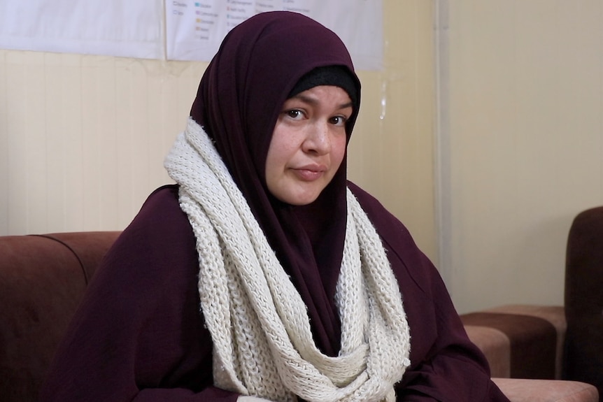 Zeinab Ahmed wears a head scarf as she sits on a couch in a room near a wall with a map pinned to it.
