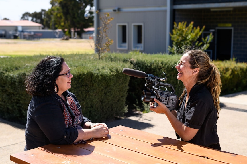 Nancy, left, smiles as she sits at an outside table while being filmed by Shalom Almond, right.