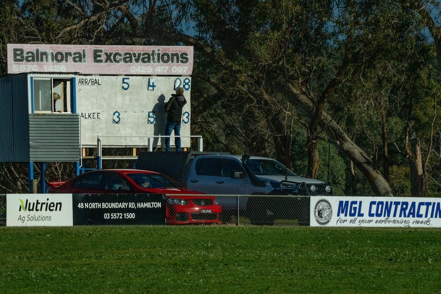 A scoreboard at a country football oval, with a man flipping the numbers.