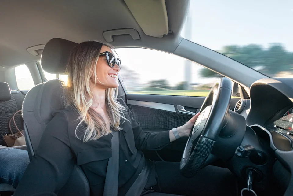 A person driving a car, smiling while looking straight ahead, wearing sunglasses and a dark outfit