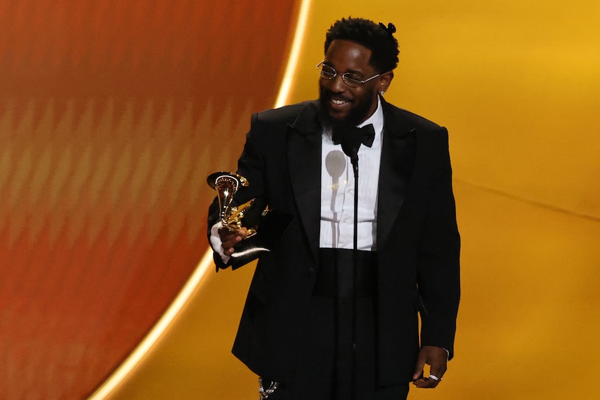 A smiling man in a tuxedo holds a statuette as he stands on a stage at an awards ceremony.