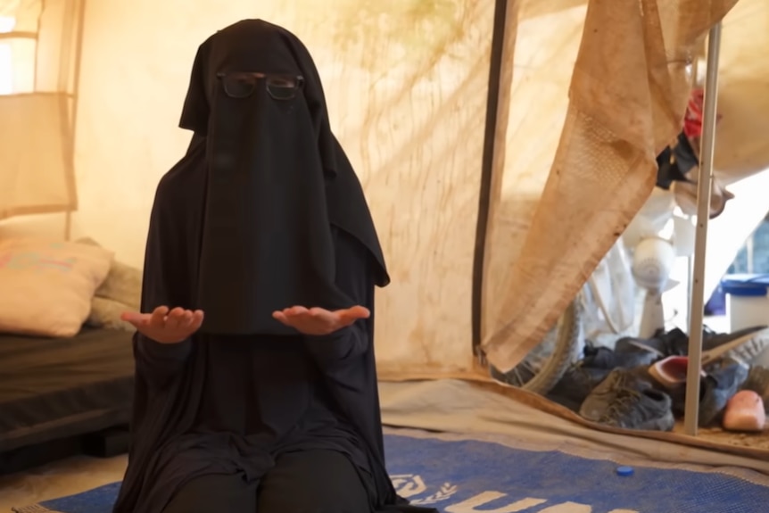 A woman wearing a black burqa sits with her hands raised in a tent.