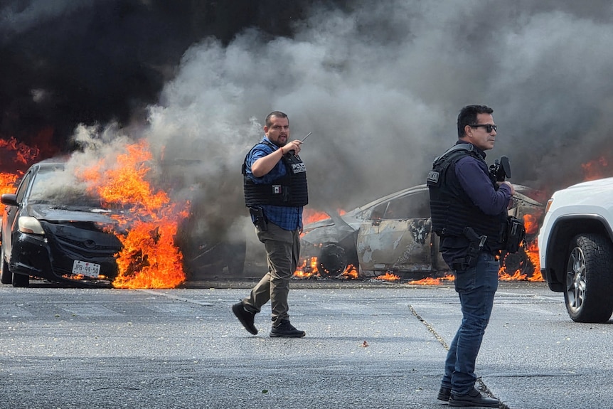 police officiers stand in front of two cars with orange flames coming from them.