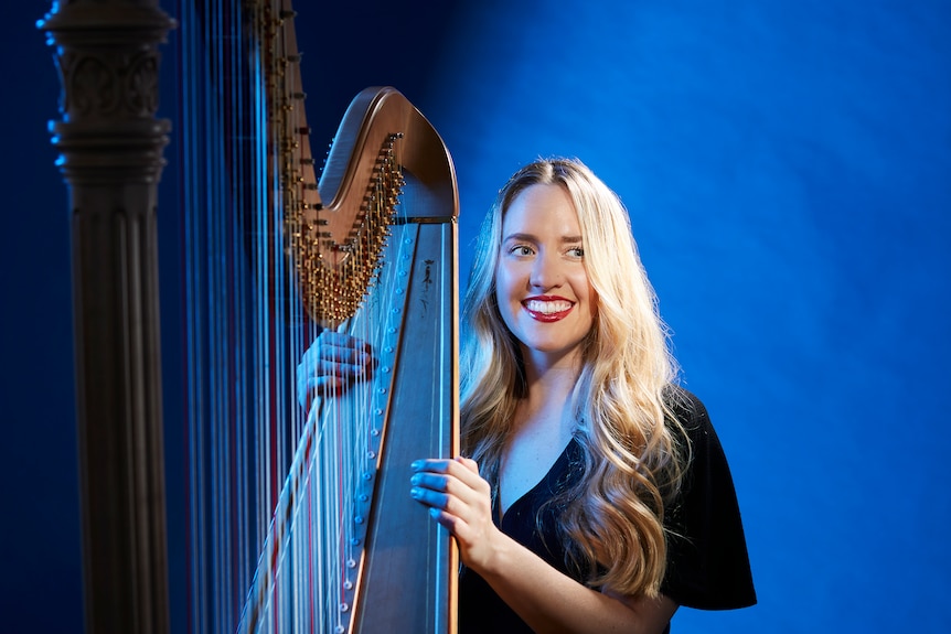 A harpist stands smiling with her harp against a blue backdrop