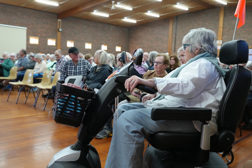 A crowd of seated people in a fall, with an older woman on a mobility scooter in the foreground.