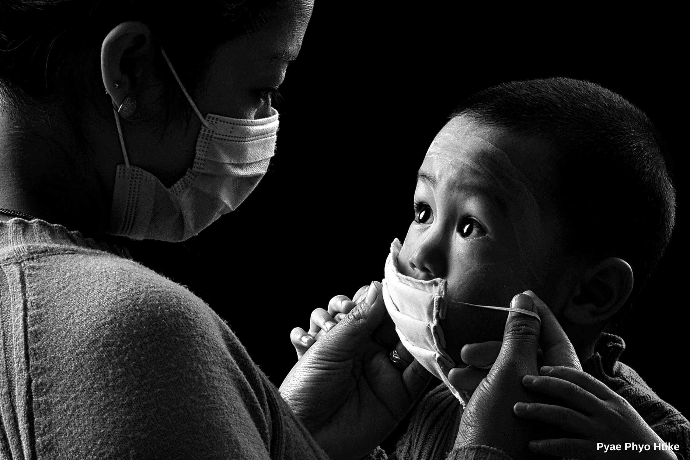 A black-and-white photo of a child looking up as an adult helps adjust their face mask. The scene conveys care and attentiveness in a pandemic context