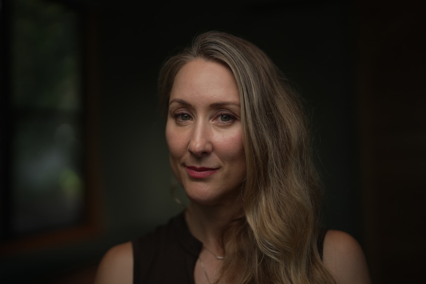 A woman with blonde hair swept to the side wearing a dark sleeveless dress sitting in a dark room.