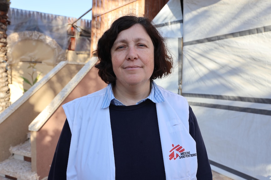 A woman wearing a vest with the MSF logo smiles slightly, in front of a tent.