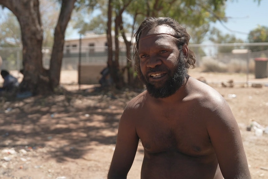 A man in a remote NT commity looks seriously at the camera. He is not wearing a shirt.