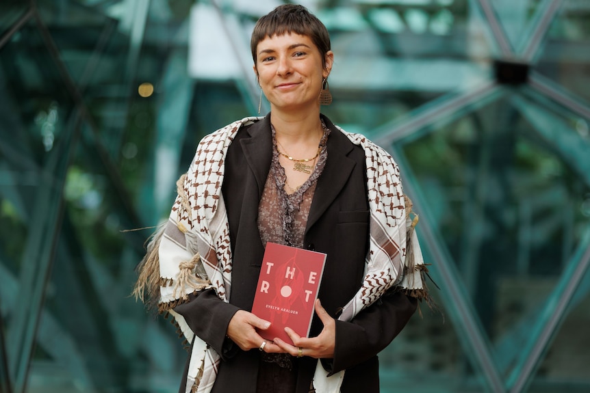 Evelyn Araluen, a smiling short-haired woman, wearing a shawl over a brown jacket, holding a book in her hands.