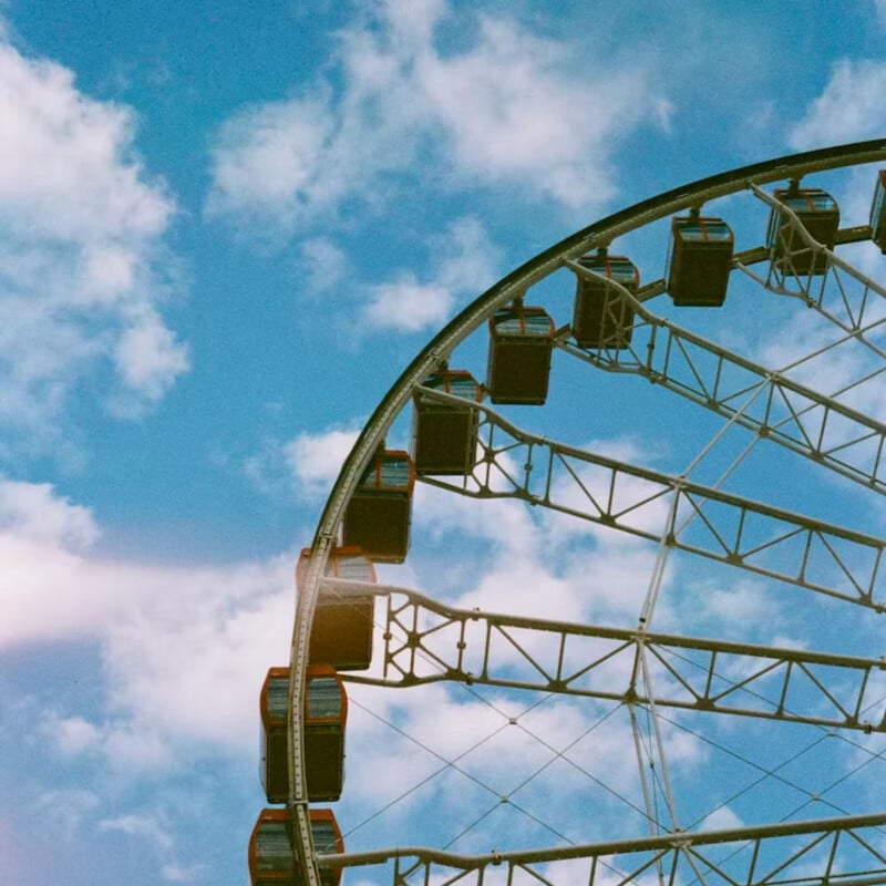 A partial view of a Ferris wheel against a bright blue sky with scattered clouds, capturing several gondolas and metal supports in sunlight.