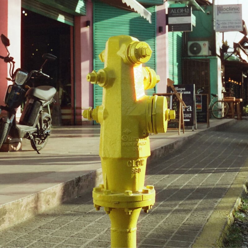 A bright yellow fire hydrant stands on a paved sidewalk next to storefronts and a parked motorcycle in a sunlit urban street scene.