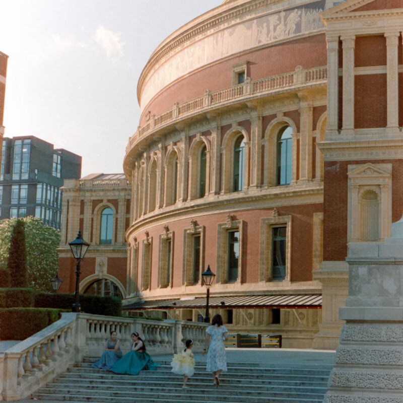 Four people in formal dresses, including a child in yellow, sit and walk on steps outside a grand, circular building with tall windows and intricate stonework, bathed in warm sunlight.