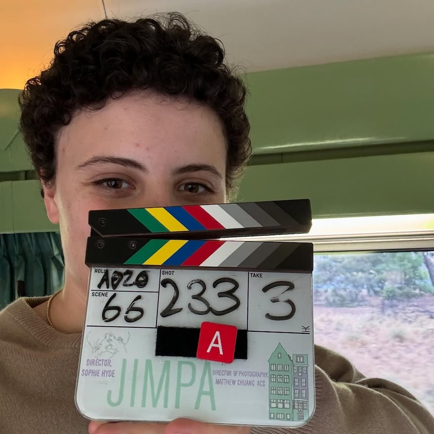 A person with short, dark, curly hair covers half their face with a movie clapper board which has numbers and the word 'Jimpa'