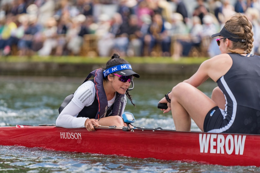 A female rowing coxswain is shouting instructions to a crew. She is wearing a visor and sunglasses.